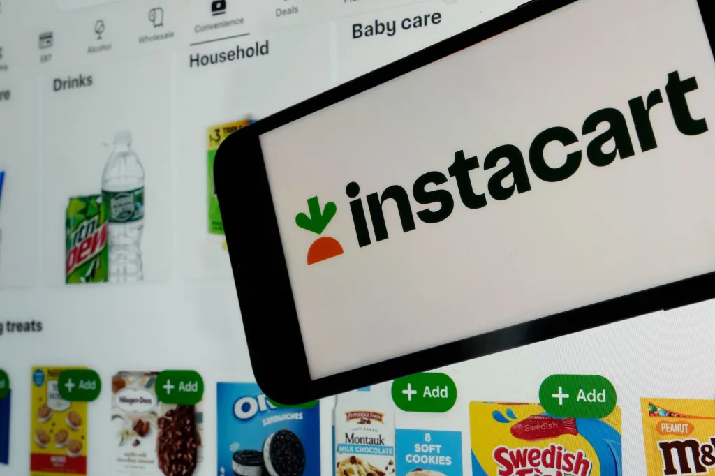 An Instacart logo on a phone screen over a webpage showing various groceries for sale.
