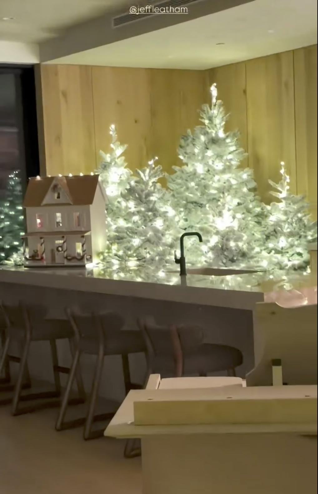 A kitchen counter with a miniature house and several frosted Christmas trees behind it, lit with warm white lights.