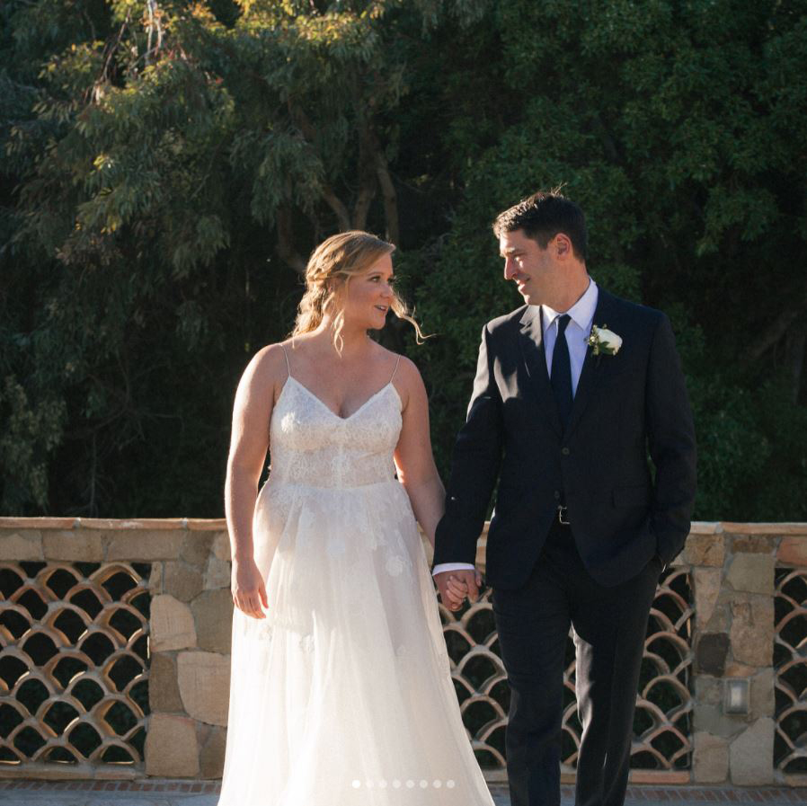 Amy Schumer, in a white flowing gown, and Chris Fischer, in a black suit and black tie, holding hands at their wedding.
