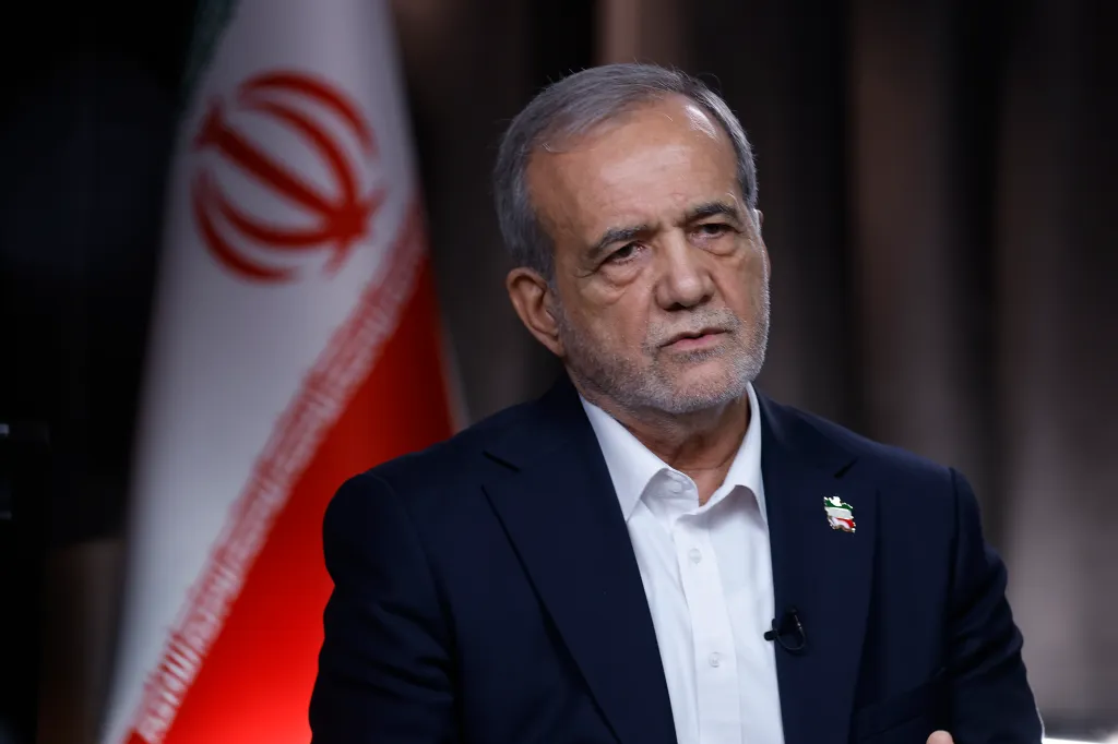 Iranian President Masoud Pezeshkian in a dark suit and white collared shirt with the Iranian flag in the background.