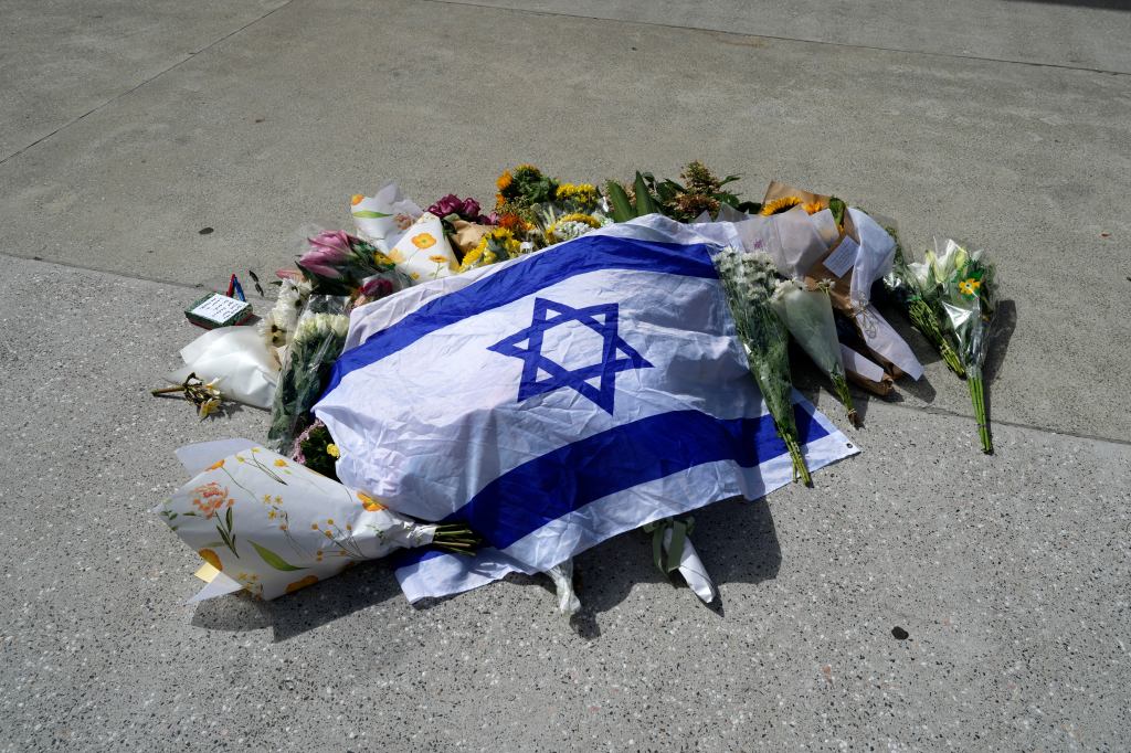 An Israeli flag laid on the ground amongst flowers, tributes, and a small notebook with pens.