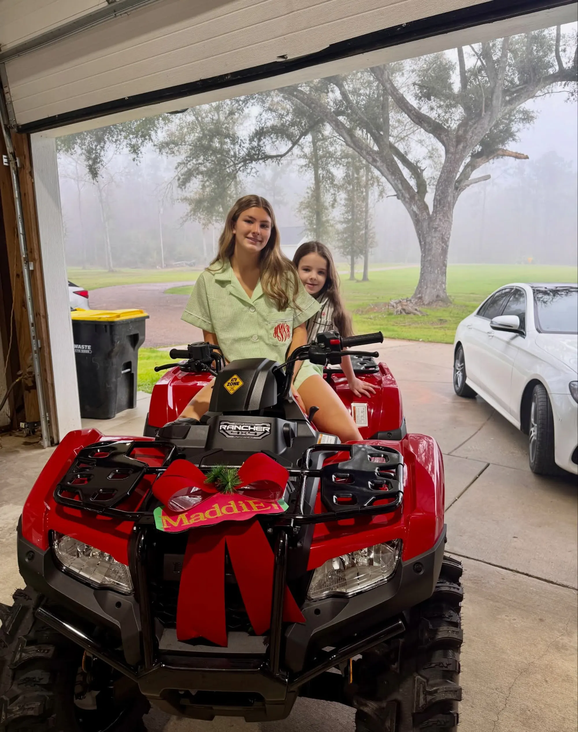 A smiling Maddie sitting atop her present, a red ATV, with her half-sister Ivey sitting behind her.