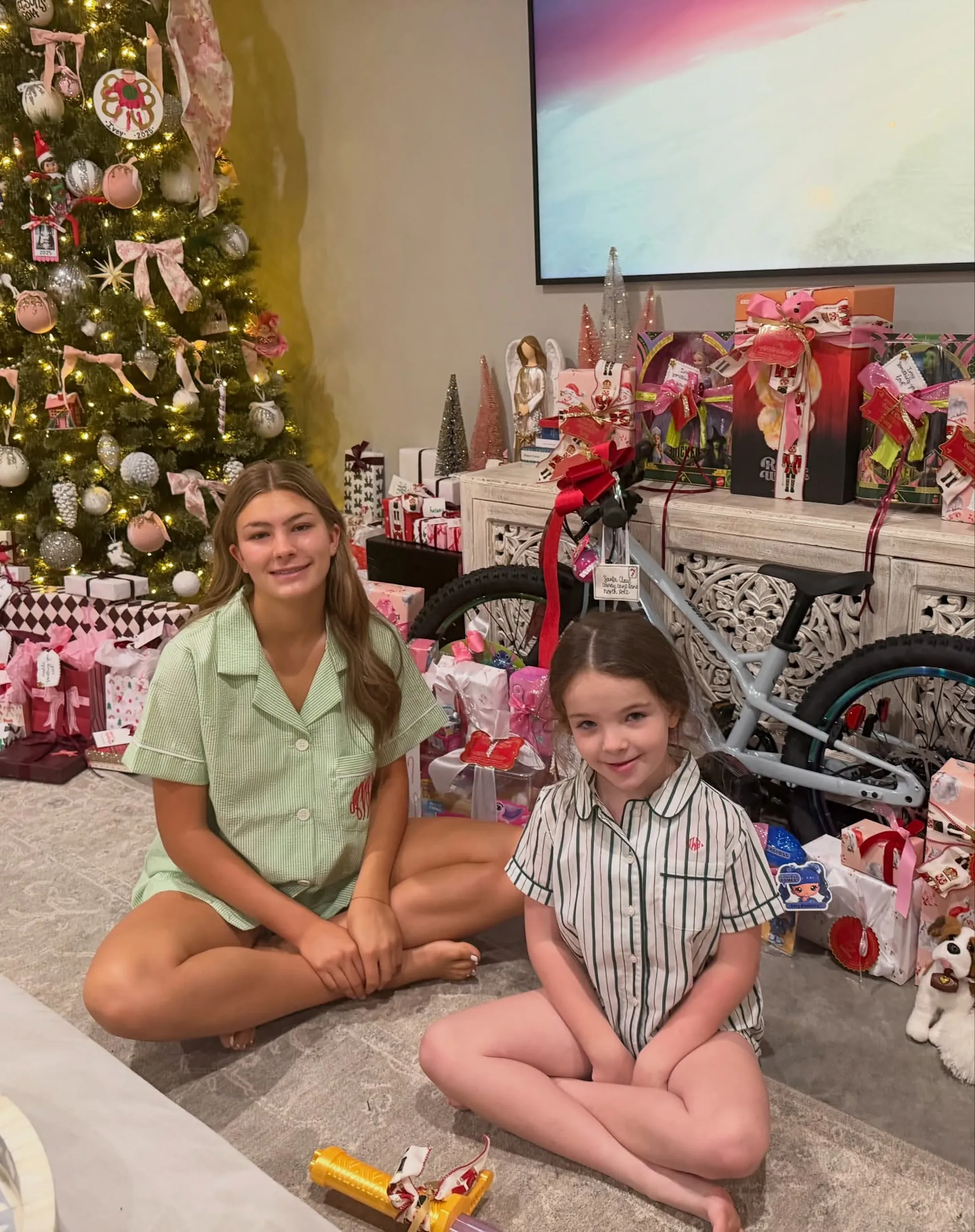 Two girls in pajamas sitting in front of a Christmas tree and gifts, including a light blue bicycle.