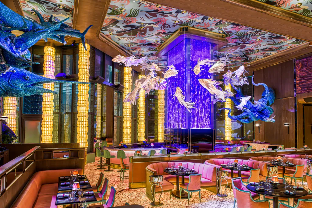 Interior of Sexy Fish Miami restaurant with a colorful, fish-themed ceiling, pink booth seating, and a glowing purple water feature.