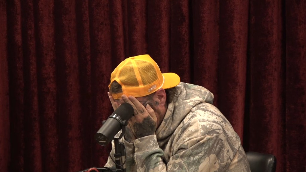 Jelly Roll with hands on his face and wearing a camouflage hoodie and yellow cap.