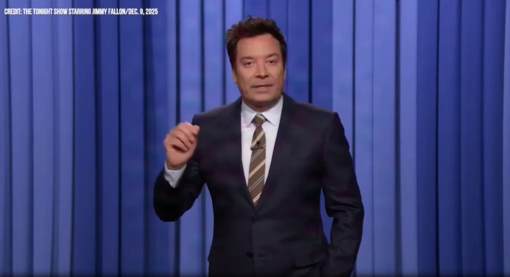 Jimmy Fallon in a suit and striped tie, gesturing with his right hand in front of a blue background.