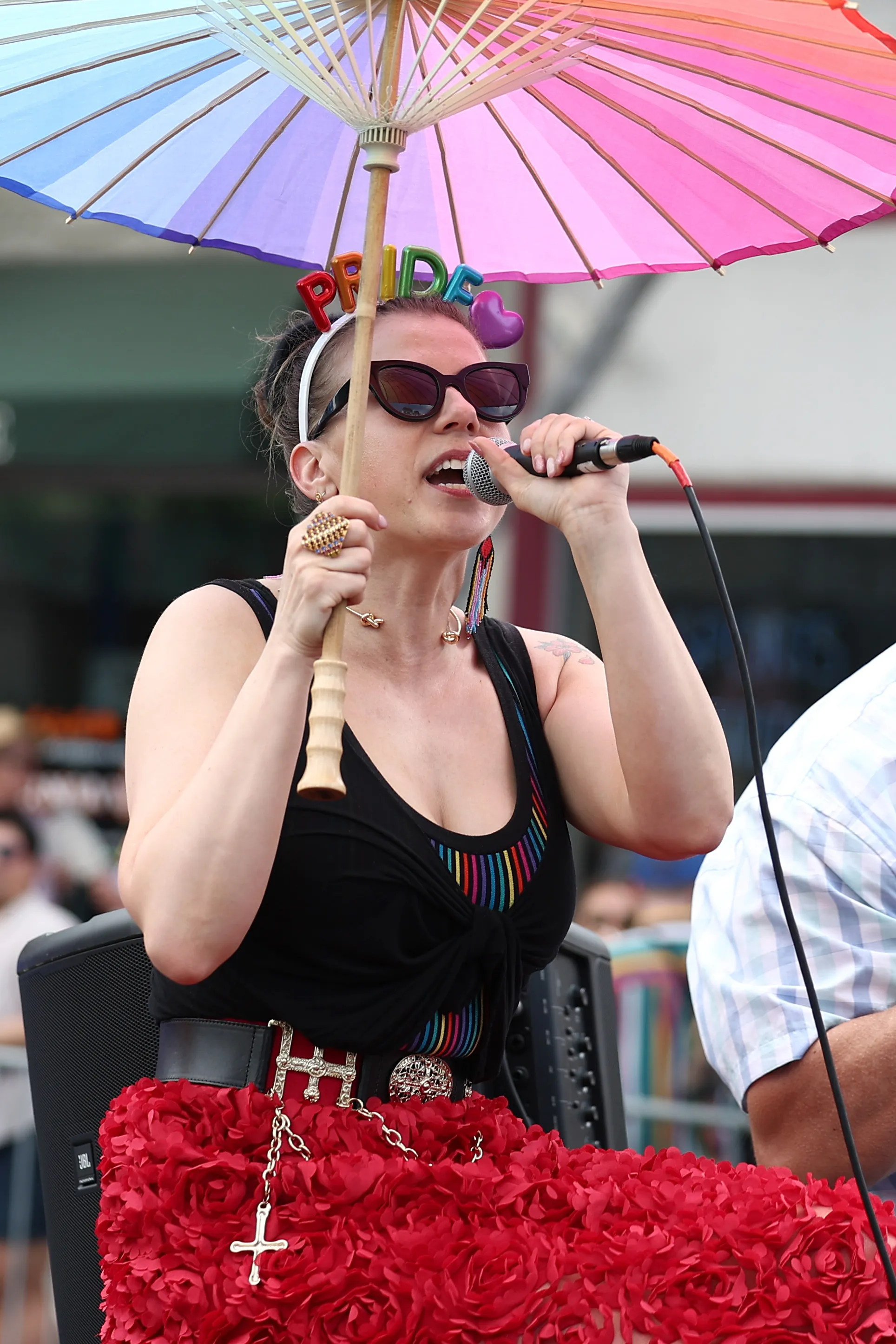 Jodie Sweetin in sunglasses, singing into a microphone, holding a rainbow parasol, and wearing a