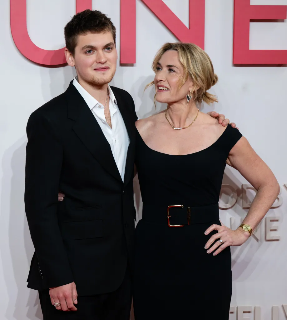 Kate Winslet in a black off-the-shoulder dress and Joe Anders in a black suit with an open white shirt at the