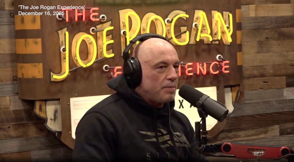 Joe Rogan wearing headphones and a hoodie sitting in front of a neon sign for 