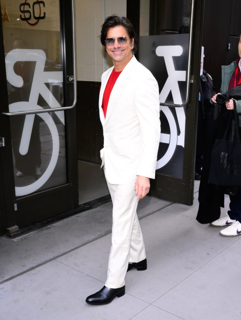 John Stamos in a white suit and red shirt, wearing sunglasses and smiling.