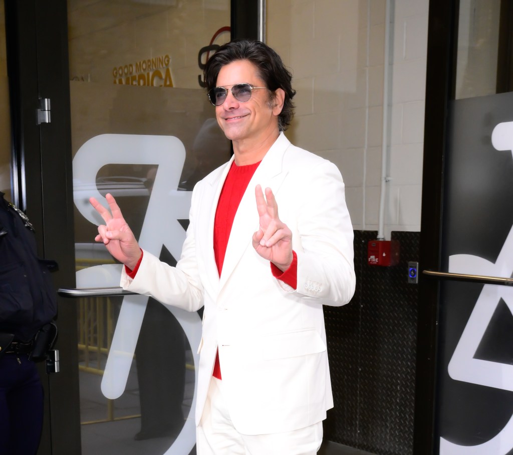 John Stamos in a white suit and red shirt makes peace signs.