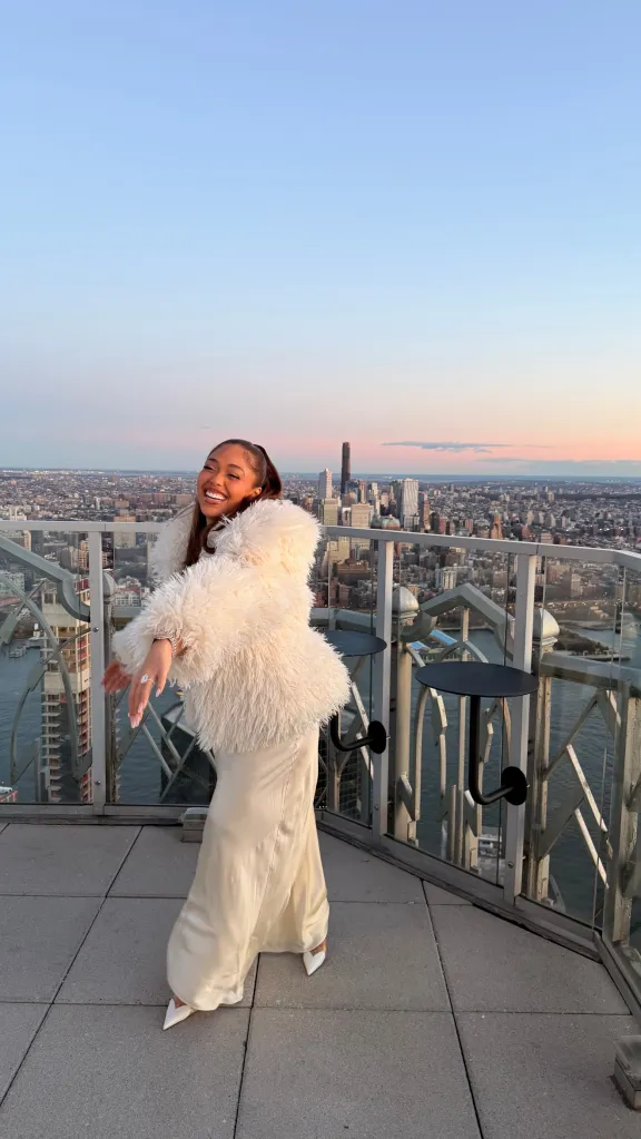 Jordyn Woods in a white fluffy coat and long satin dress posing on a rooftop with a city view at sunset.