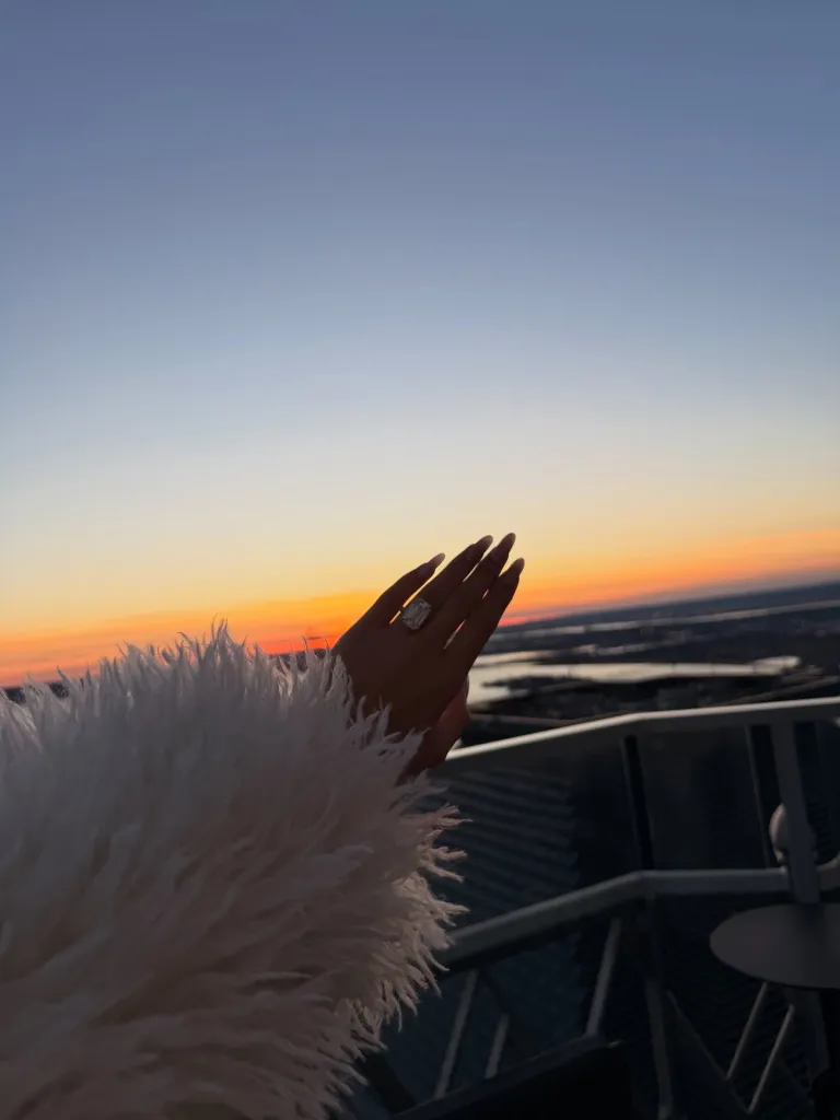 Jordyn Woods' hand with a large ring, raised towards a sunset sky.