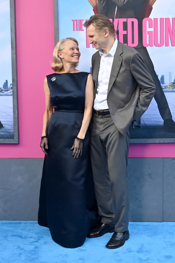 Pamela Anderson in a navy gown and Liam Neeson in a gray suit smile at each other on a red carpet.
