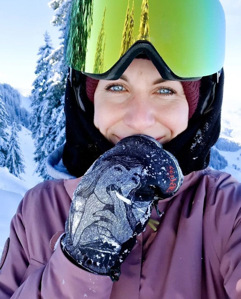 A woman in ski gear smiling with a snowy mountain and trees in the background.