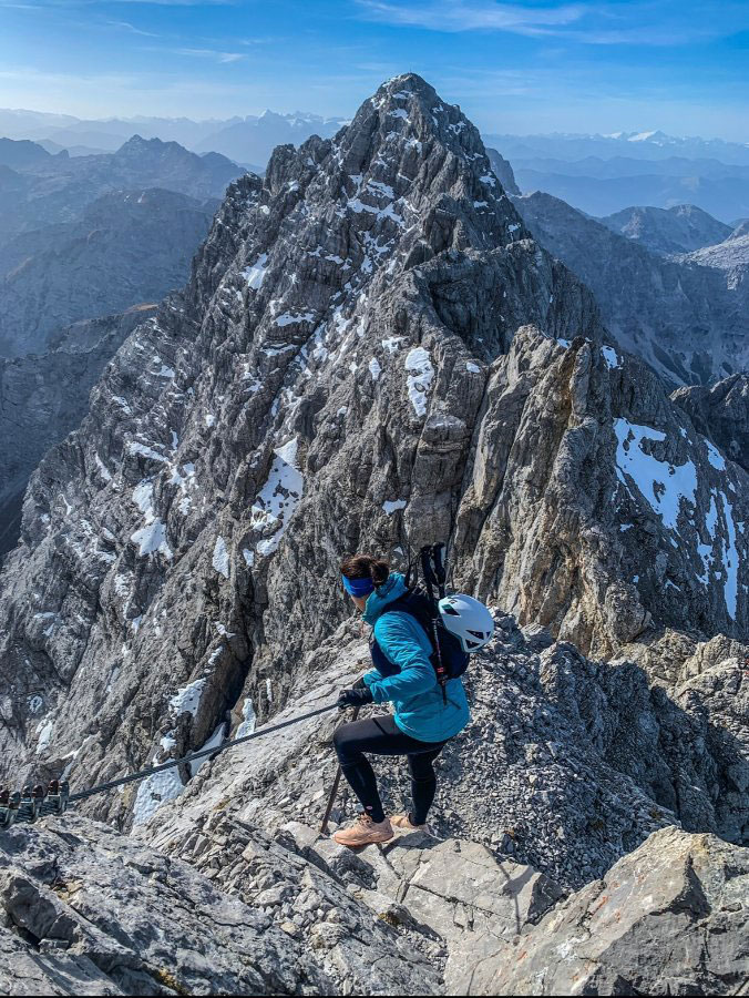A person climbing a rocky mountain with snow patches, secured by a cable.