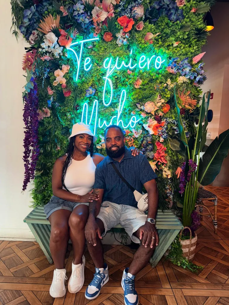 Kandi Burruss and Todd Tucker sitting on a bench in front of a floral wall with a 