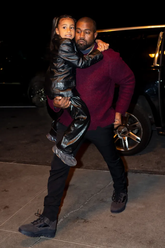 Kanye West holding North West, both wearing dark clothing, outside at night.