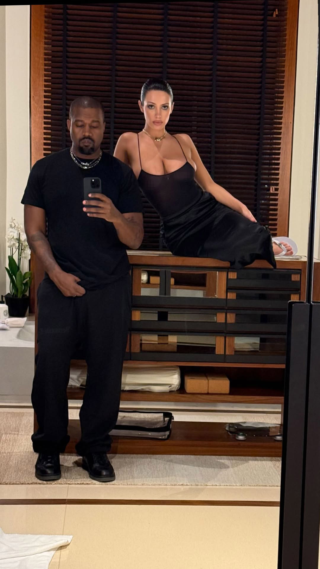 Kanye West taking a mirror selfie with Bianca Censori sitting on a wooden cabinet.