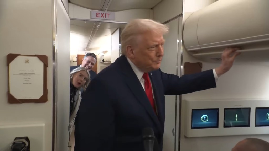 Donald Trump in a suit and red tie stands on Air Force One, reaching up with his right arm, as Karoline Leavitt peaks out a bathroom door behind him with a man smiling over her shoulder.