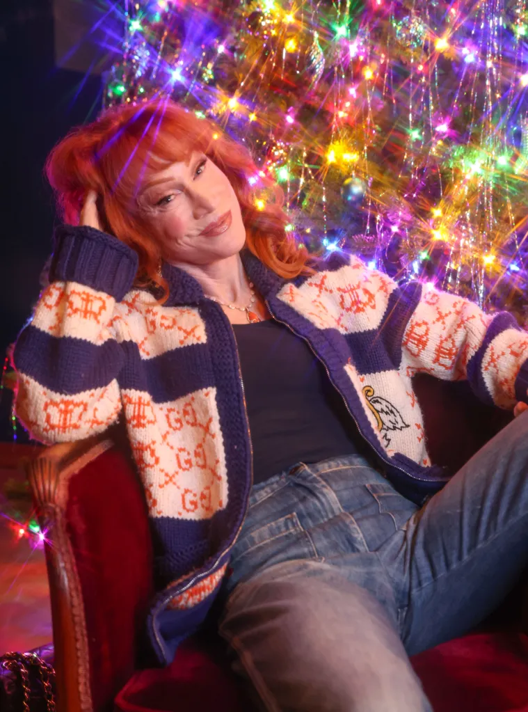 Kathy Griffin posing in a blue and white patterned sweater and jeans, with a brightly lit Christmas tree in the background.