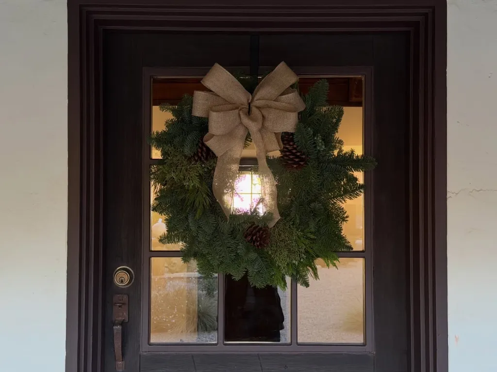 Christmas wreath with burlap bow and pinecones hanging on a dark wood door.