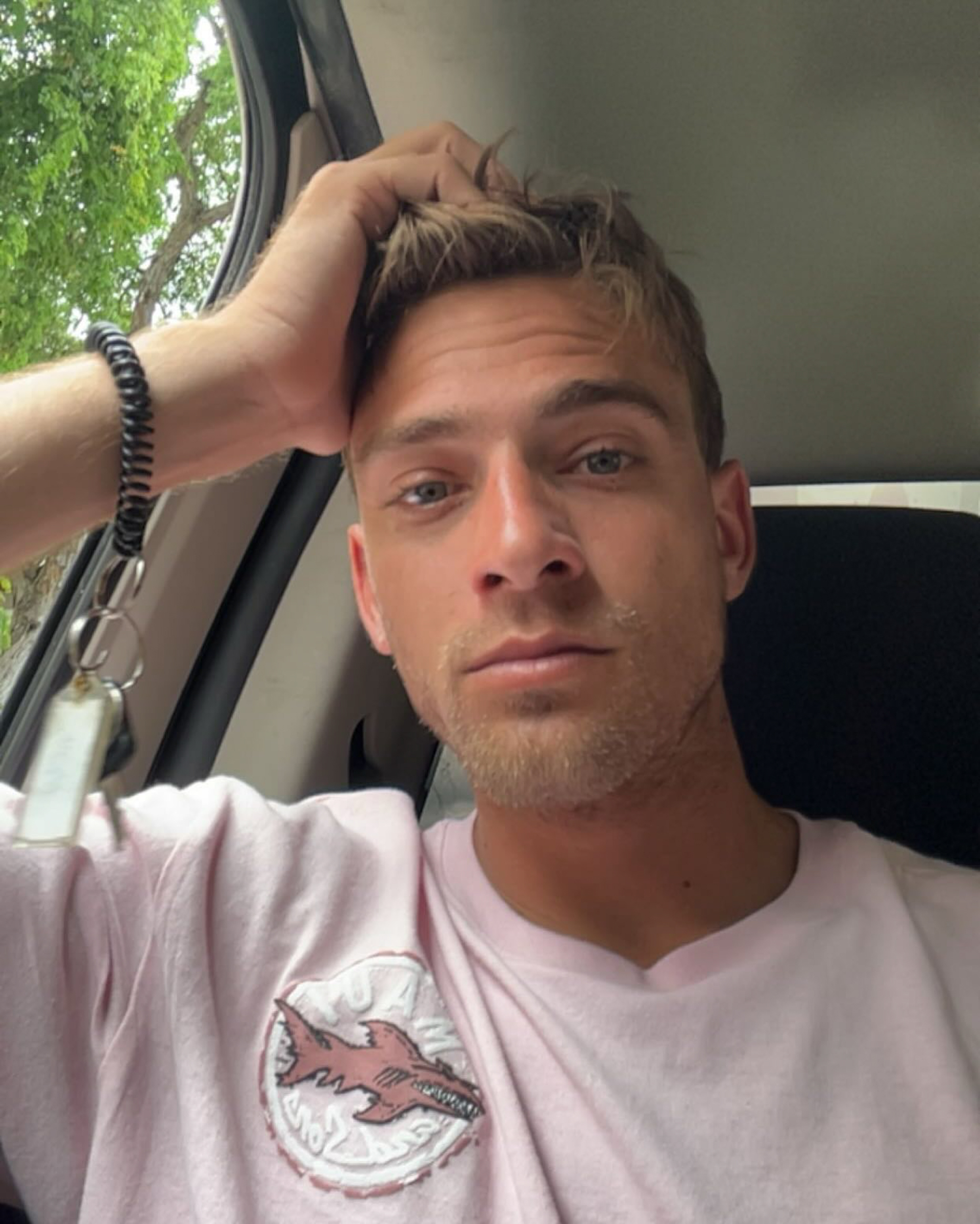 A man in a pink t-shirt with a shark logo, holding keys with a coiled bracelet on his wrist, looks directly into the camera.