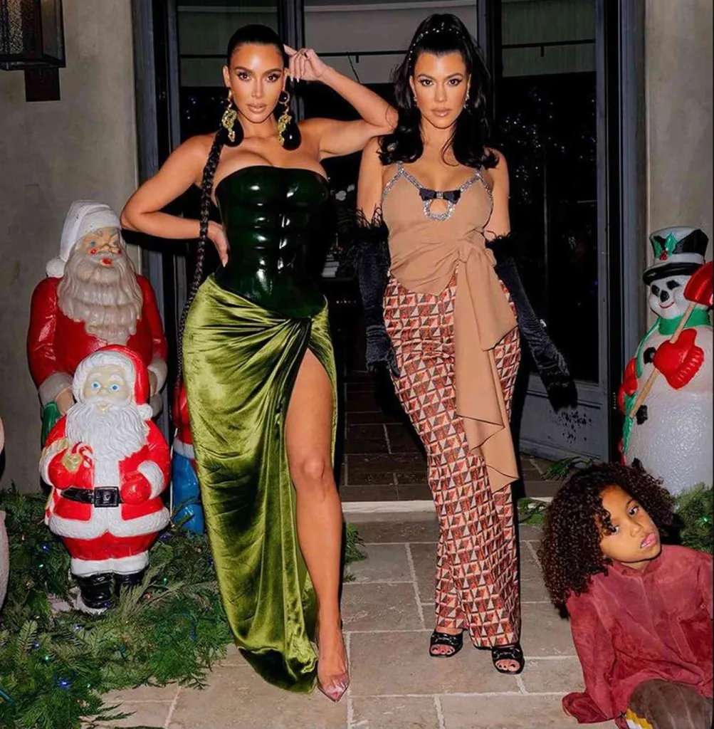Kim and Kourtney Kardashian standing outside with Christmas decorations, with a child crouching in the foreground.