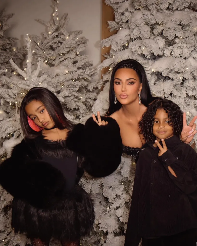 Kim Kardashian and her two children posing in front of white flocked Christmas trees.