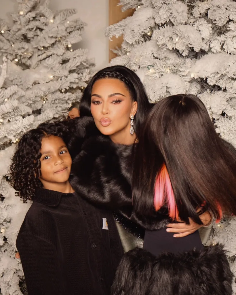 Kim Kardashian with two children in front of snow-covered Christmas trees.