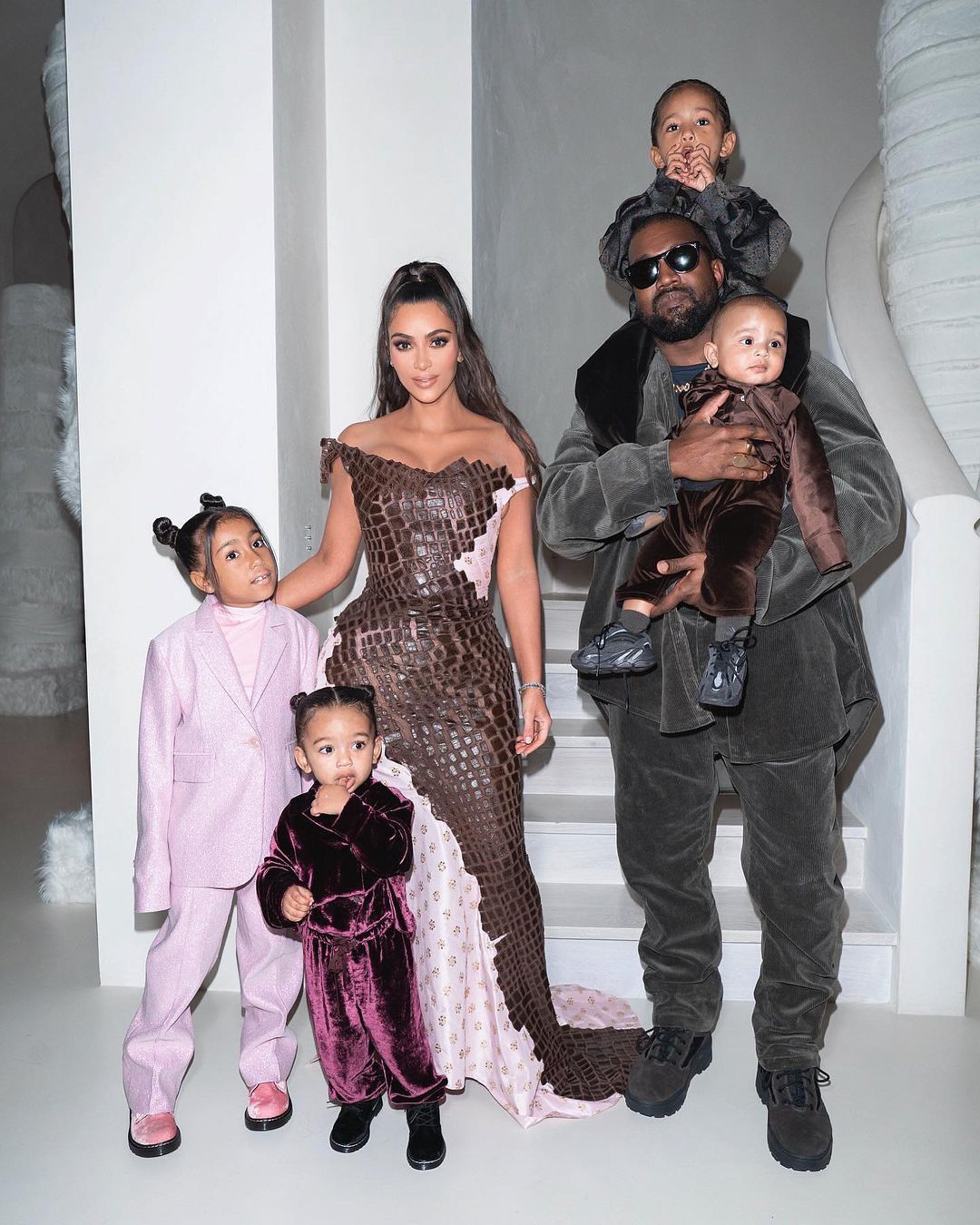 Kim Kardashian, Kanye West, and their four children stand together on a white floor.