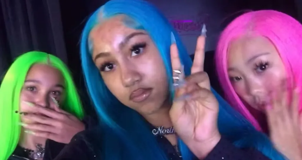 Three girls in bright wigs, one with blue hair making a peace sign, another with green hair covering her mouth, and the third with pink hair.