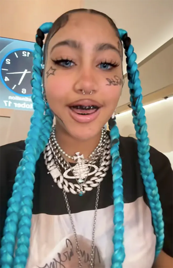 A person with bright blue braids, blue eyes, and faux face tattoos, wearing several necklaces and a nose piercing.