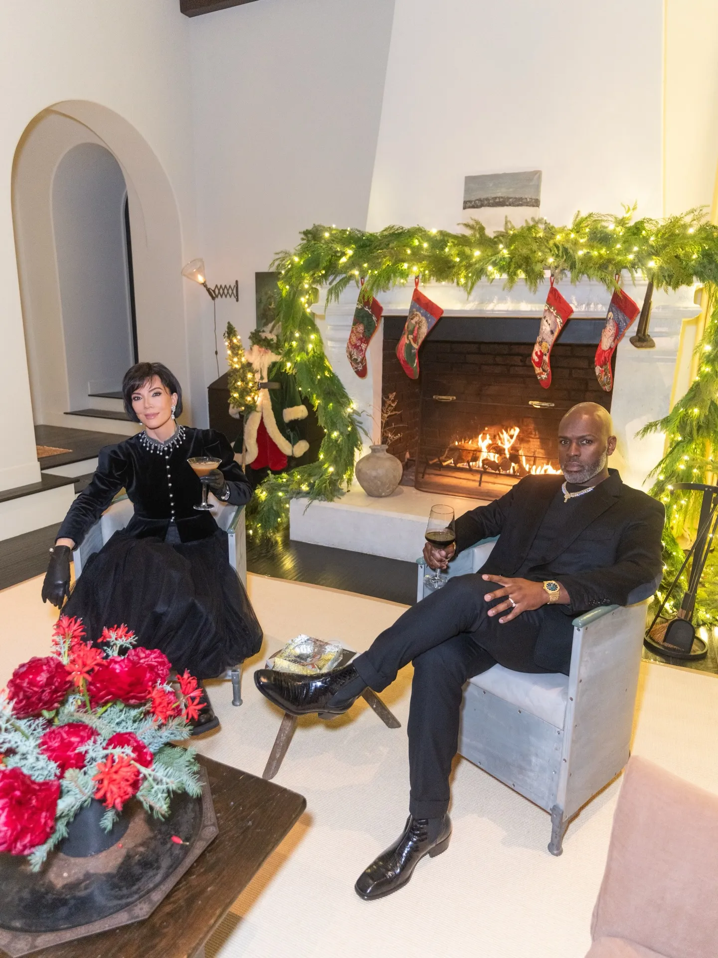 Kris Jenner and Corey Gamble seated in front of a fireplace decorated with Christmas garland and stockings.