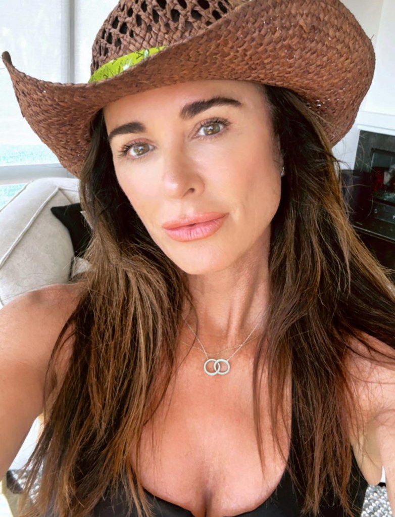 Kyle Richards posing while wearing a brown straw hat and a silver necklace with two interlocking rings.