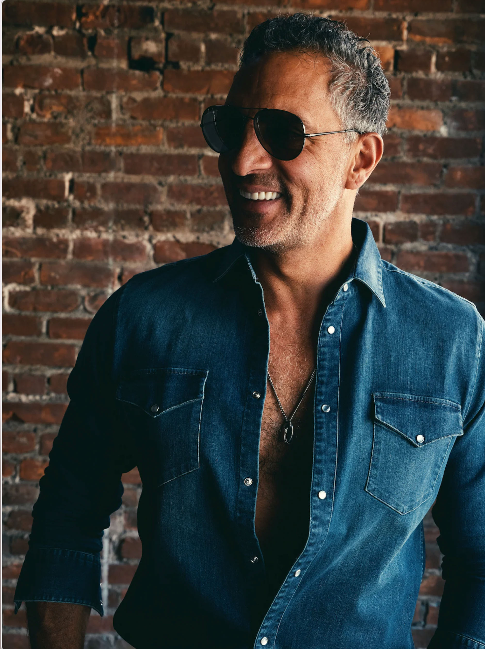 Mauricio Umansky smiling in sunglasses and an unbuttoned denim shirt.