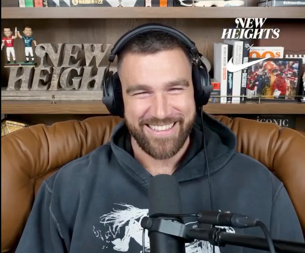 Travis Kelce laughing while wearing headphones and a hoodie.