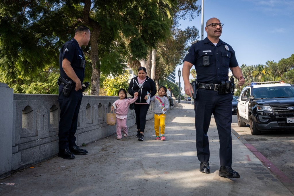 Police officers standing on a sidewalk near a woman and two young girls, with a police vehicle behind them.