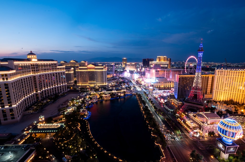 Aerial view of the Las Vegas Strip at sunset with neon lights, casinos, and a lake in the foreground.