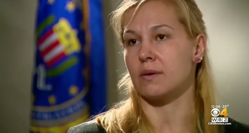 A light-skinned woman with blonde hair, Laura Schwartzenberger, looks right while a blue flag with the FBI seal is in the blurred background.