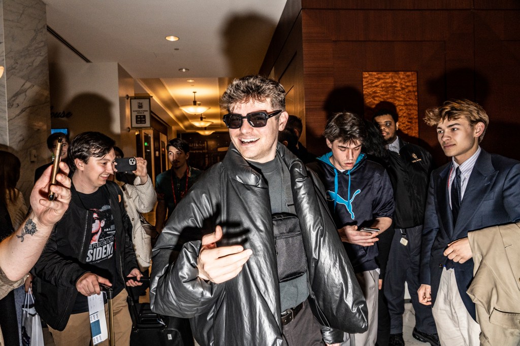 Nicholas Fuentes, an American white supremacist, smiling while walking through a hallway, surrounded by a crowd of people.