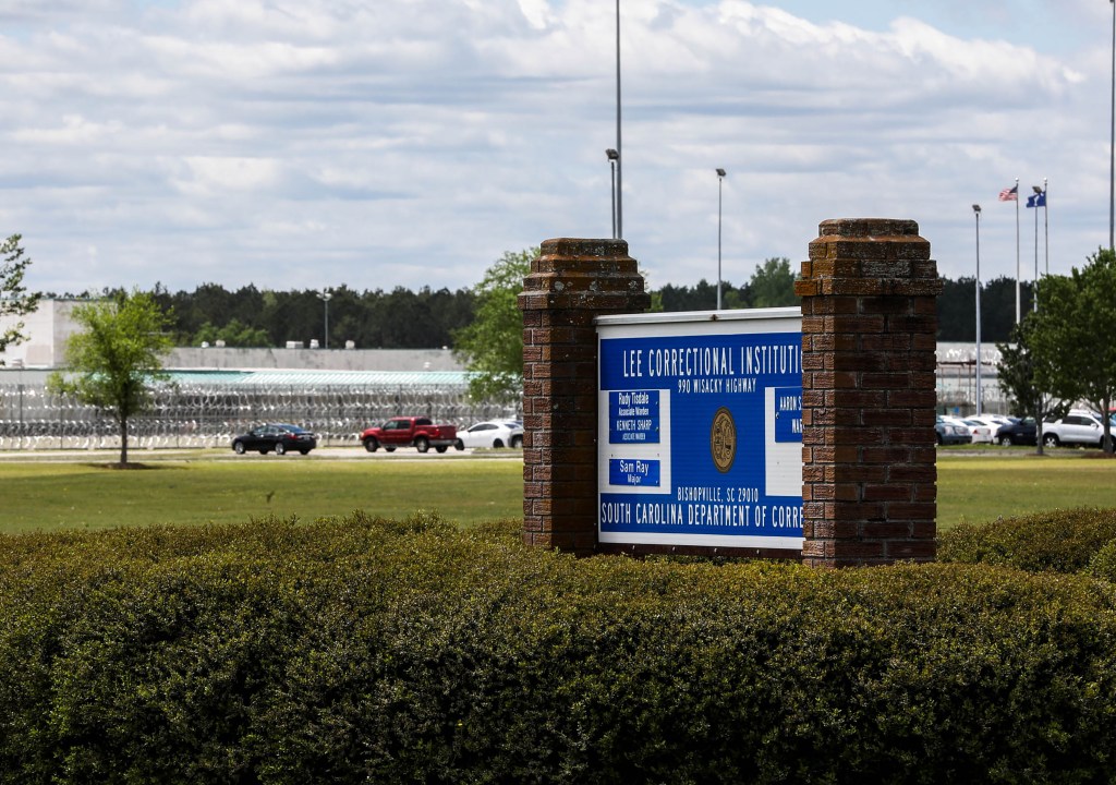 Sign for Lee Correctional Institution with a prison fence and vehicles in the background.