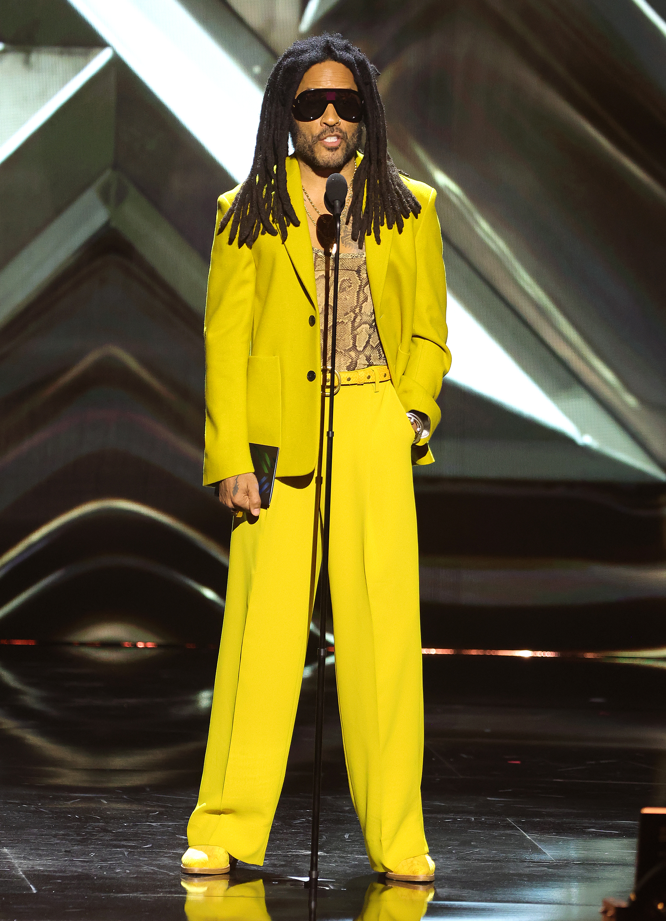 Lenny Kravitz on stage wearing a yellow suit with a snakeskin-patterned shirt and sunglasses.