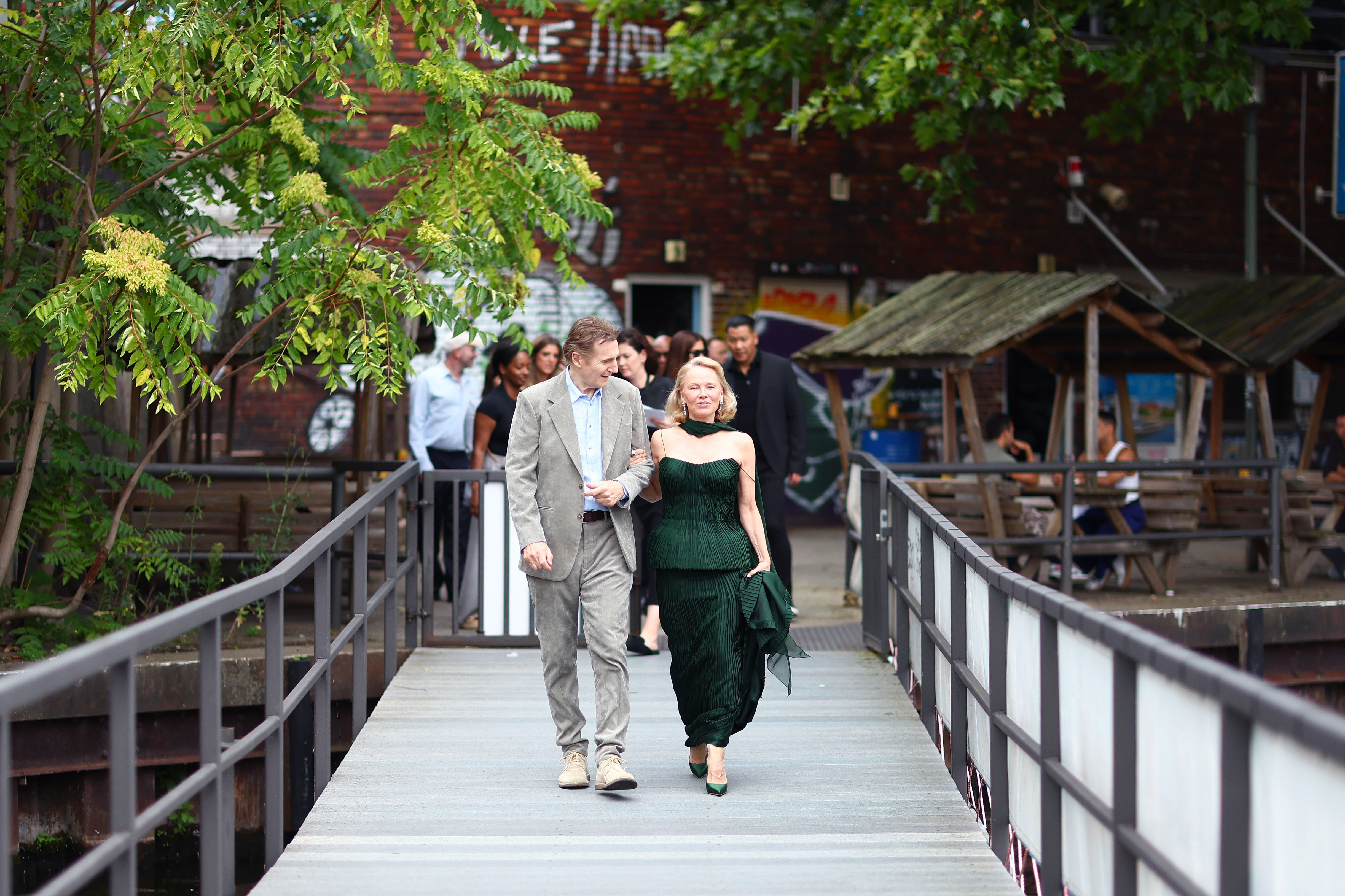Liam Neeson and Pamela Anderson walking arm-in-arm on a boardwalk.