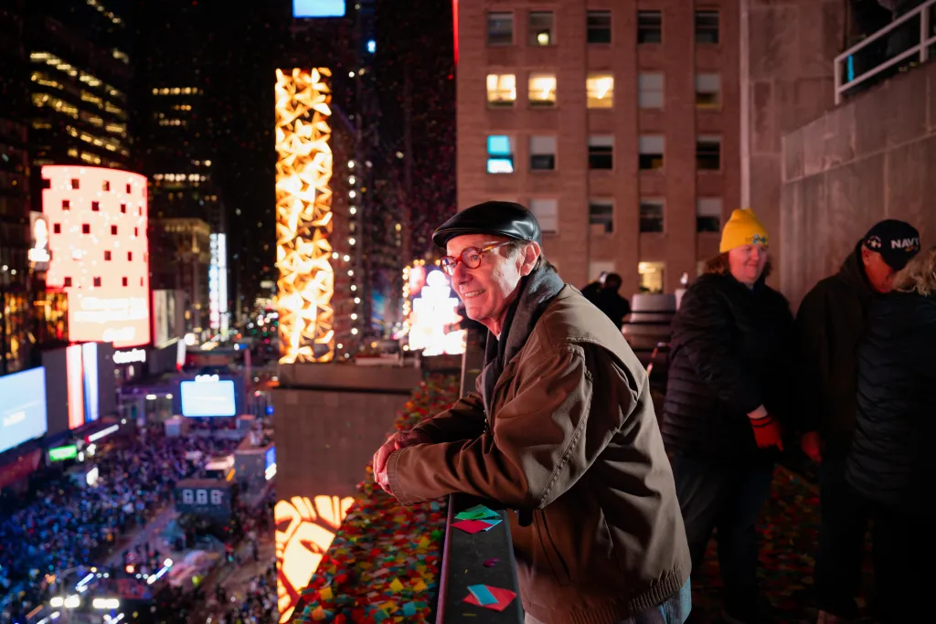 Treb Heining, also known as the Confetti King, stands on a balcony overlooking Times Square during New Year's Eve, with confetti on the railing in front of him.