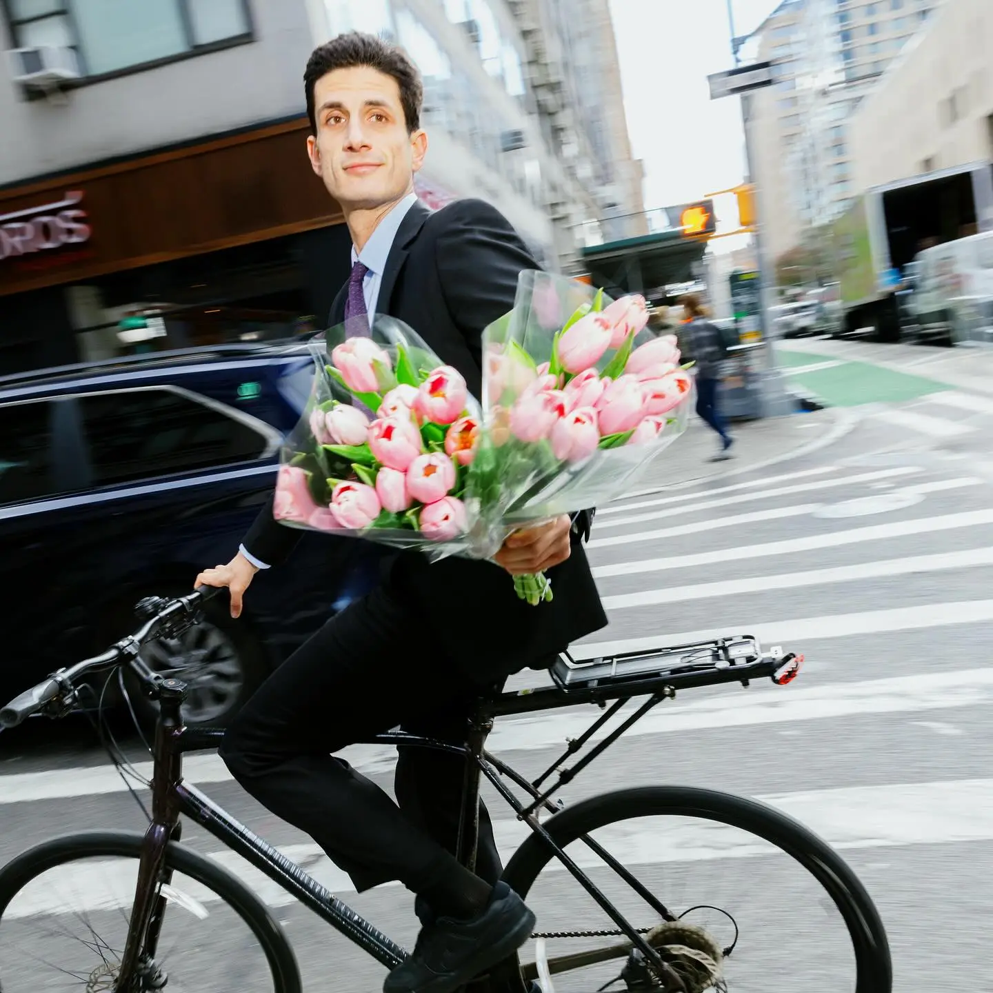 Jack Schlossberg riding a bicycle in a suit while carrying two large bouquets of pink tulips.
