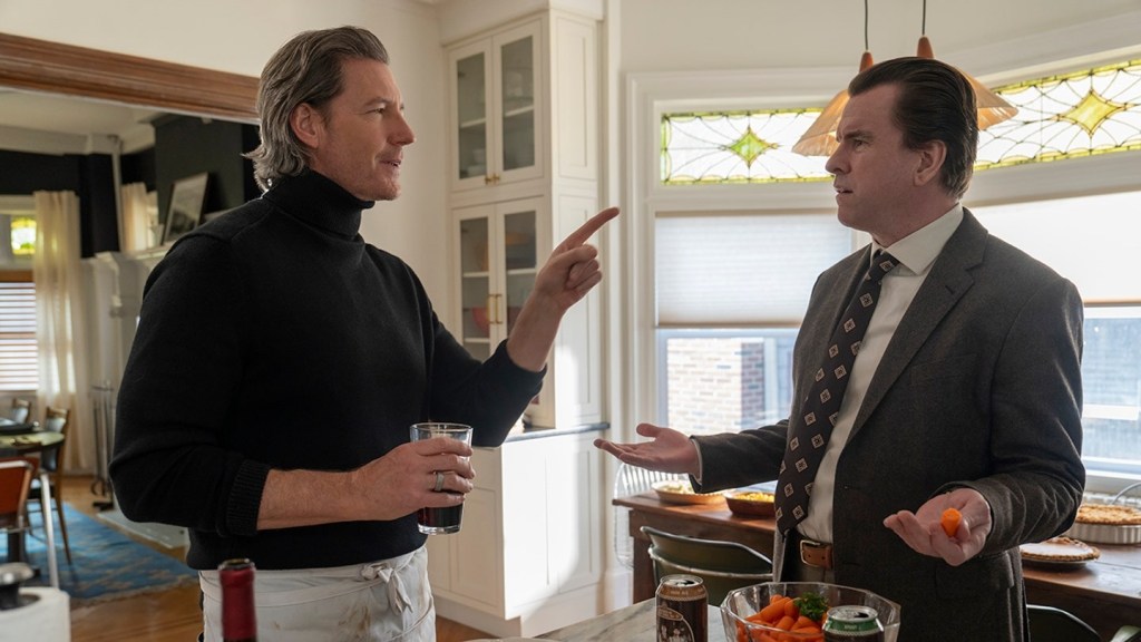 Two men, one in a black turtleneck and one in a suit, talking in a kitchen.