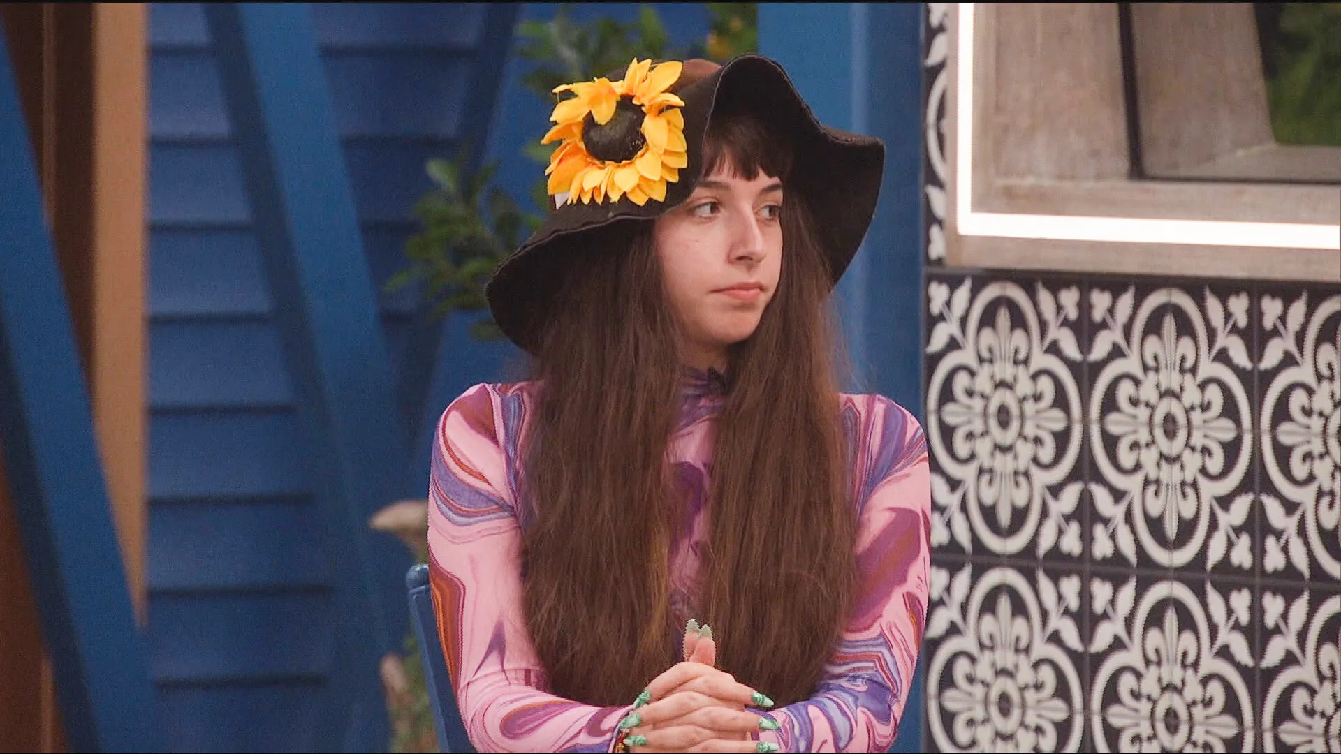 Ava Pearl, a woman with long brown hair, wearing a brown hat with a sunflower and a purple marbled long-sleeve shirt, looks away with a serious expression.