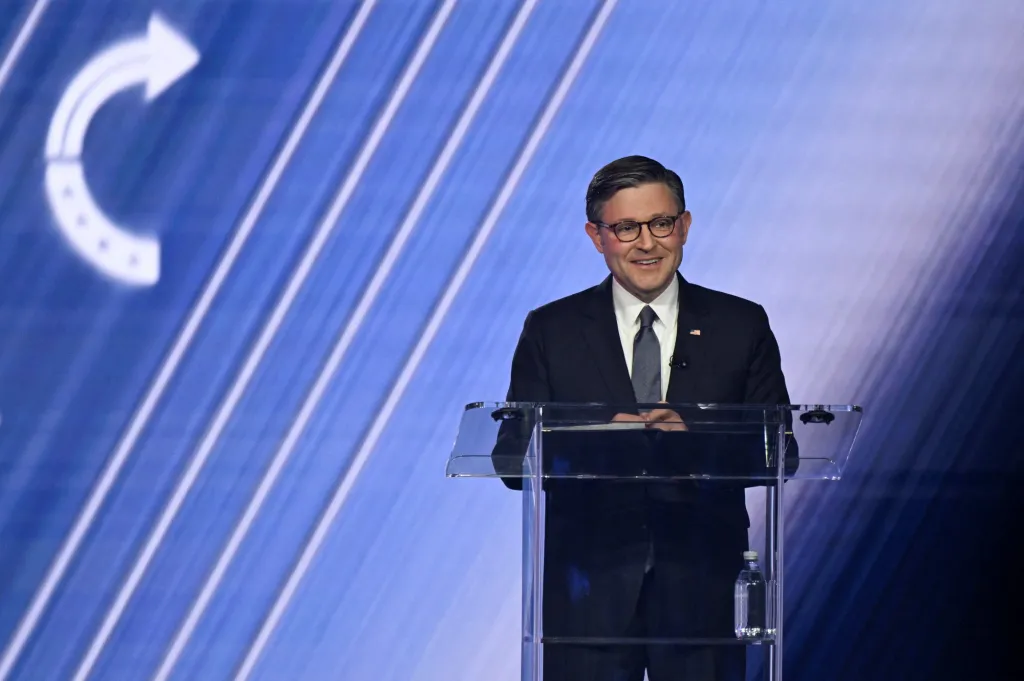 U.S. House Speaker Mike Johnson smiling at a podium during AmericaFest.