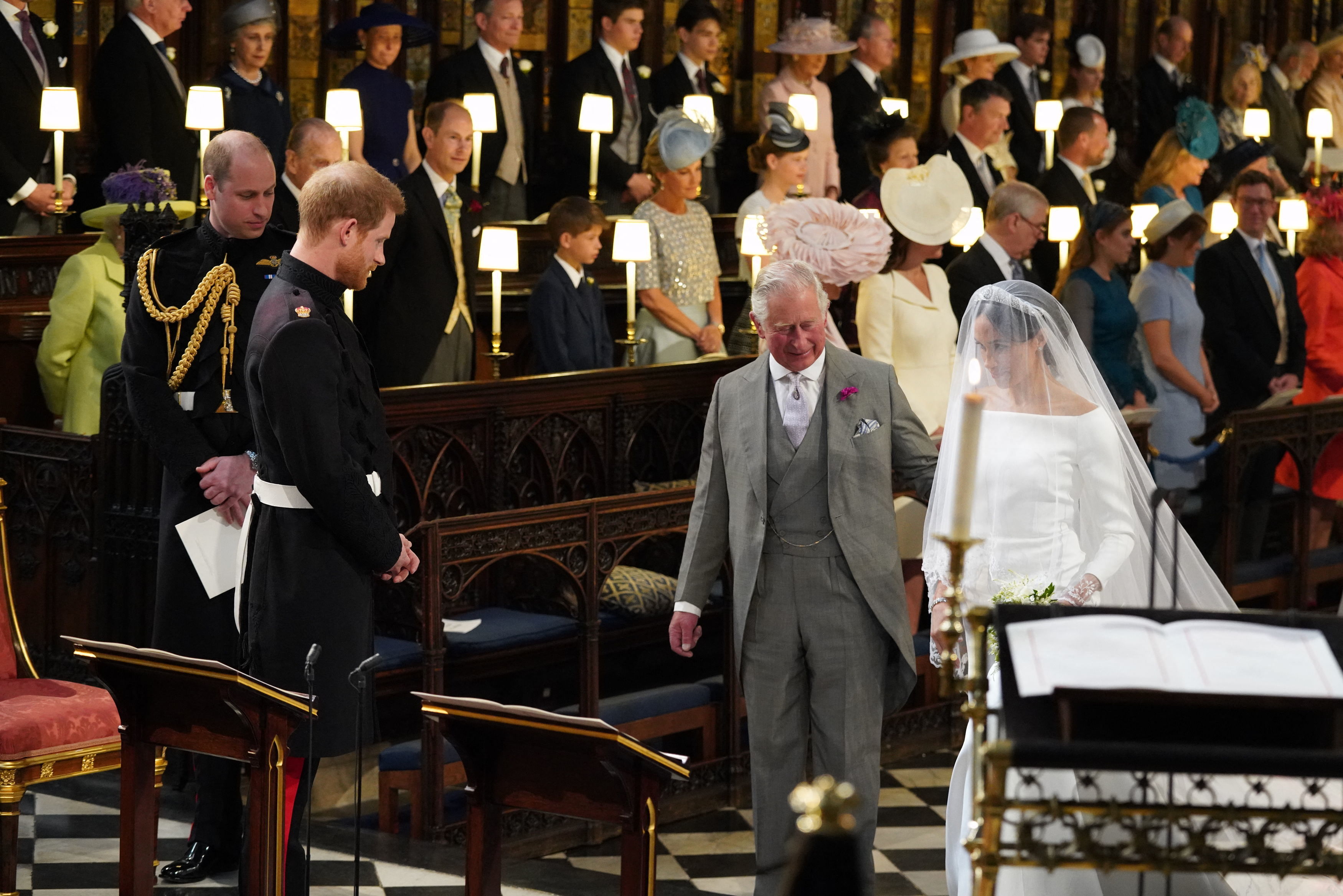 King Charles walking Meghan Markle down the aisle at her wedding to Prince Harry, with wedding guests in the background.